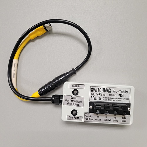 A SWITCHMAX Relay Test Box connected to a black and yellow cable, showing the input ports and set switches on a white background."