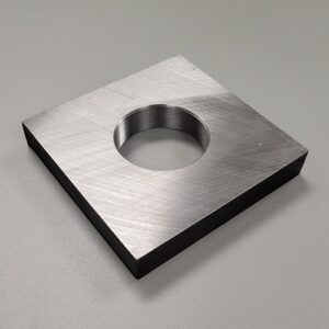 A square metal plate with a threaded central hole, resting on a rough concrete surface.