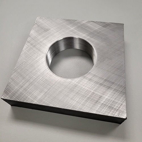 A square metal plate with a threaded central hole, resting on a rough concrete surface.