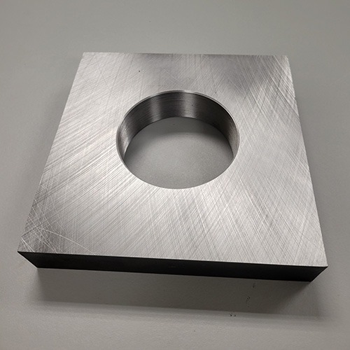 A square metal plate with a threaded central hole, resting on a rough concrete surface.