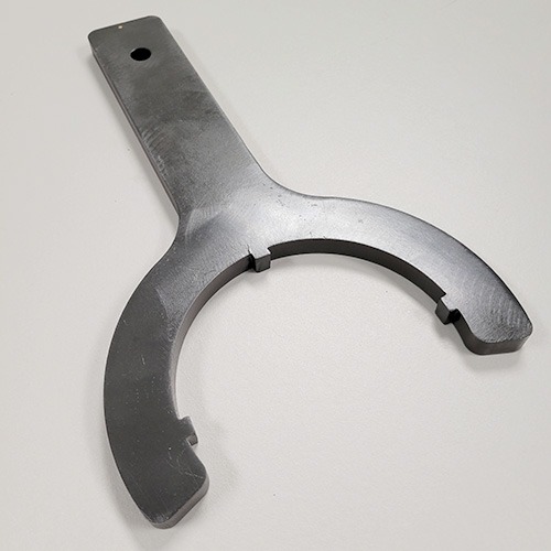 A black wrench tool with a rounded opening and flat handle, designed for a specific application, lying on a grey surface.