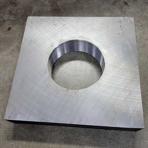 A square metal plate with a threaded central hole, resting on a rough concrete surface.